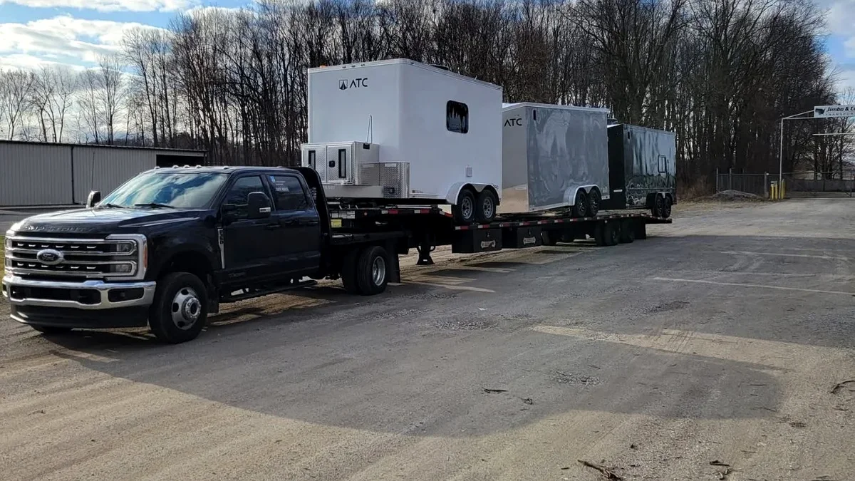 A black dually pickup pulling a step-deck flatbed loaded with three cargo trailers